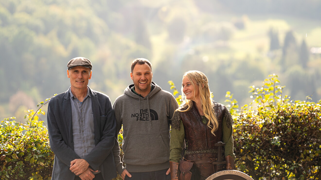Zwei Männer und eine Frau lachen in die Kamera. Im Hintergrund ist eine grüne Hecke.