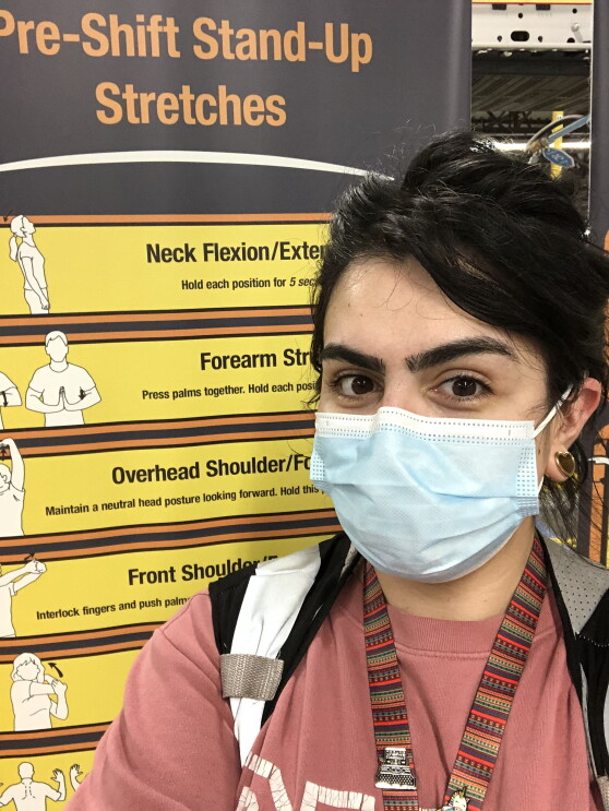 Beka Perez, an Amazon associate, smiles for a photo in front of a sign at an Amazon fulfillment center. The sign shows useful stretches for fulfillment center employees. Beka is wearing a face mask.