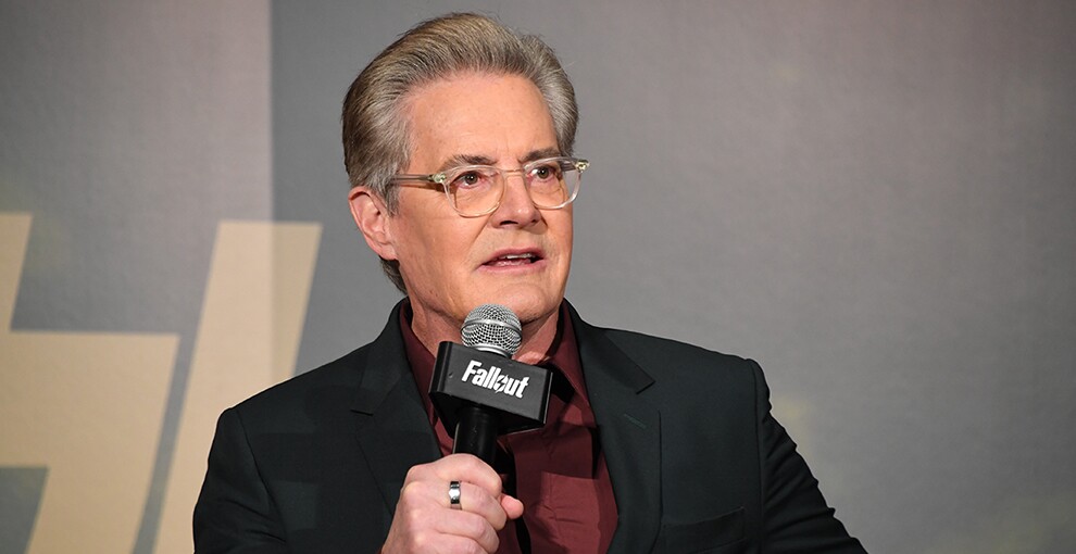 グレーの壁を背にマイクを持って話す男性。黒いジャケットで暗い紅色のシャツを着用。クリアフレームの眼鏡をかけている。