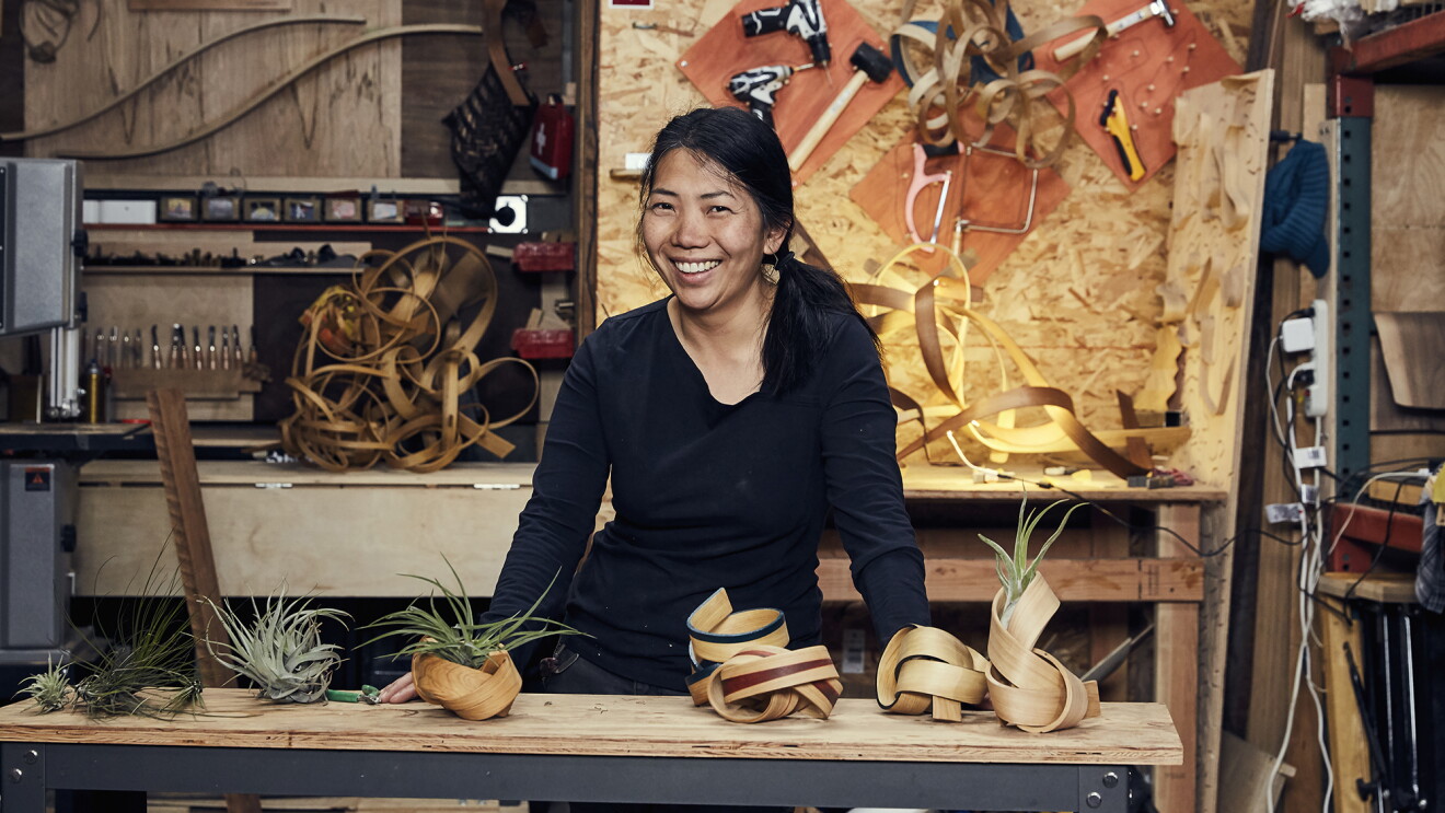 A woman stands behind a work table, smiling at the camera. In front of her are several handmade bentwood air plant containers, behind her sits more wood and tools. 