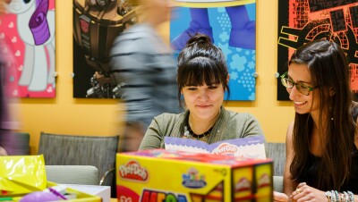 Two women in the foreground sit at a table surrounded by packaging. Other people pass by in the background. The wall has posters of My Little Pony, Play-Doh, and other Hasbro brands.