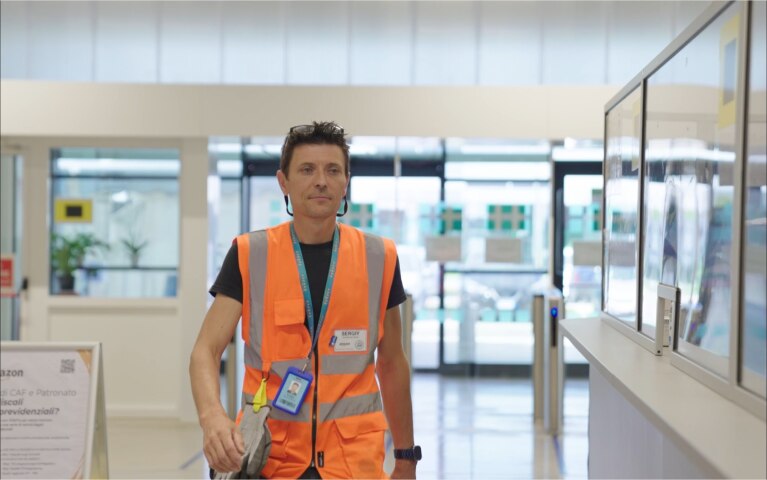 Mezzo busto di un dipendente Amazon che sorride verso la camera. Indossa un giubbino di sicurezza arancione