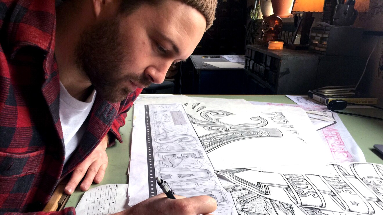 Kyler Martz in his Seattle studio, drawing the word Seattle onto a piece of sketch paper. He is wearing a red and black checked flannel shirt.