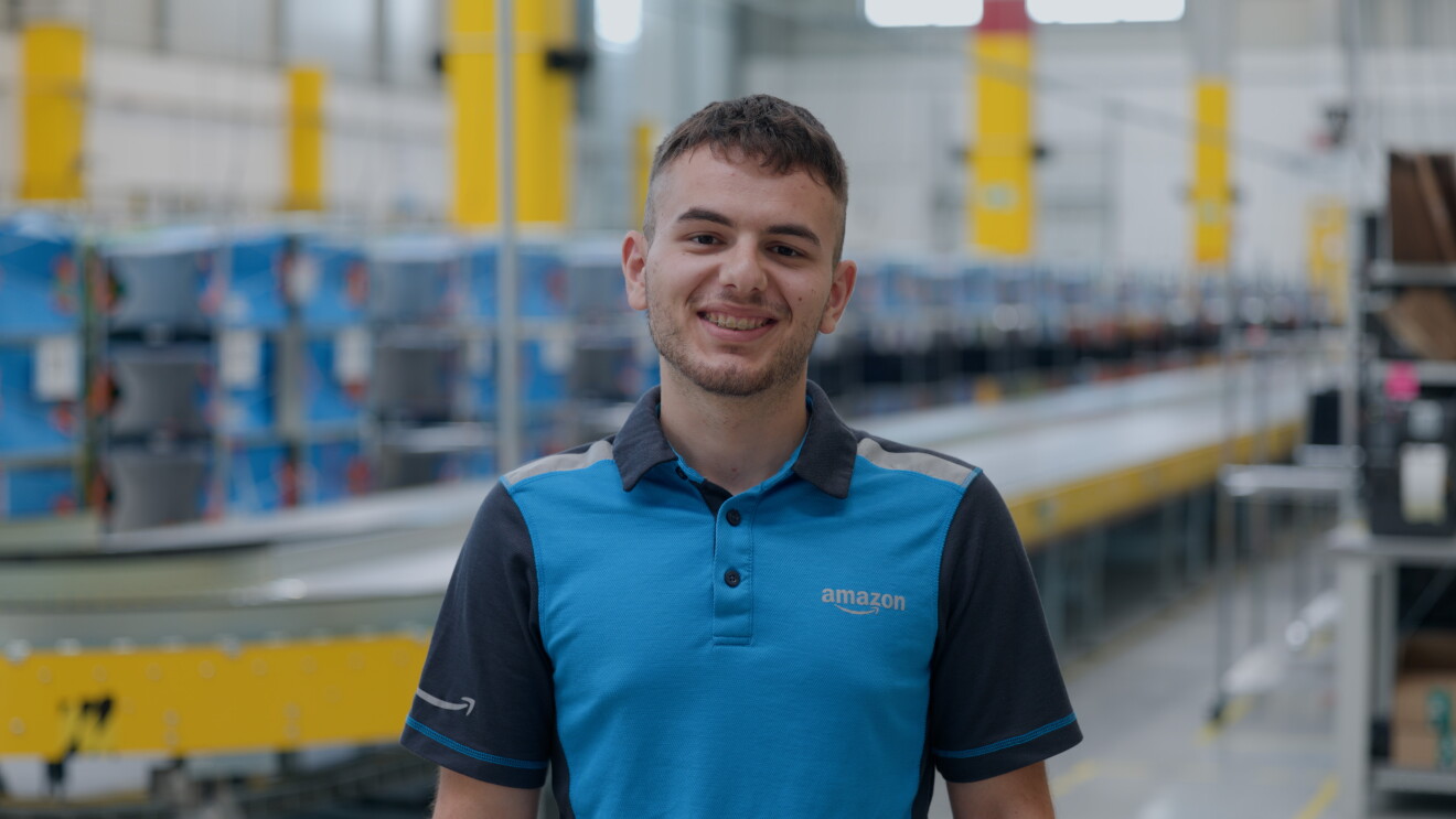 Ritratto a mezzo busto di un dipendente Amazon. Indossa una t-shirt celeste con il logo Amazon sul lato sinistro. Si trova all'interno di un magazzino e sullo sfondo c'è un rullo trasportatore