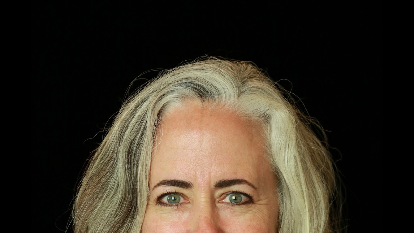 A woman with blonde hair smiles at the camera, she sits in front of a black background.