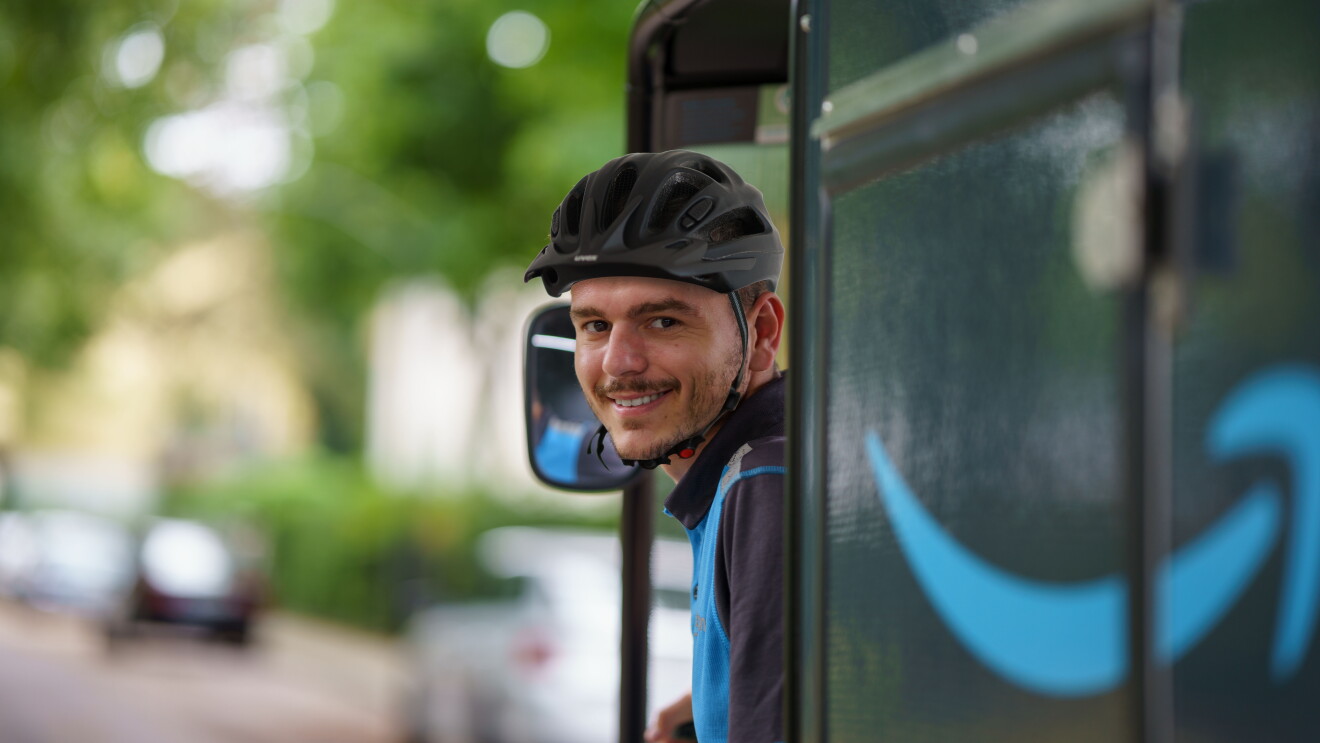 Ein junger Amazon E-Cargo-Bike-Fahrer mit schwarzem Helm und blauem Shirt auf dem E-Cargo-Bike. Er blickt sich um und lächelt in die Kamera. Verschwommen sieht man das seitlich angebrachte Amazon-Logo.