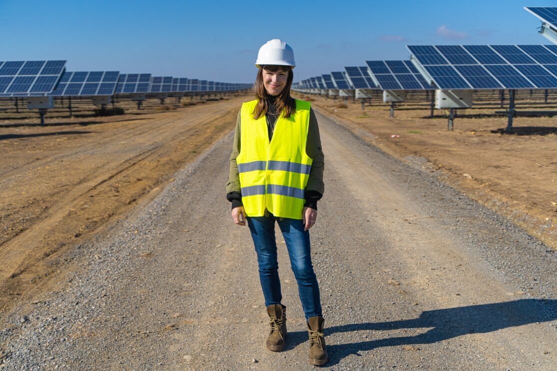 Denise Hugo, jefa de proyectos de Energías Renovables de Amazon. Ella está en medio de la foto, en un camino de tierra y entre dos líneas de placas solartes. Va vestida con unas botas marrones, unos vaqueros, casco blanco y chaleco amarillo.