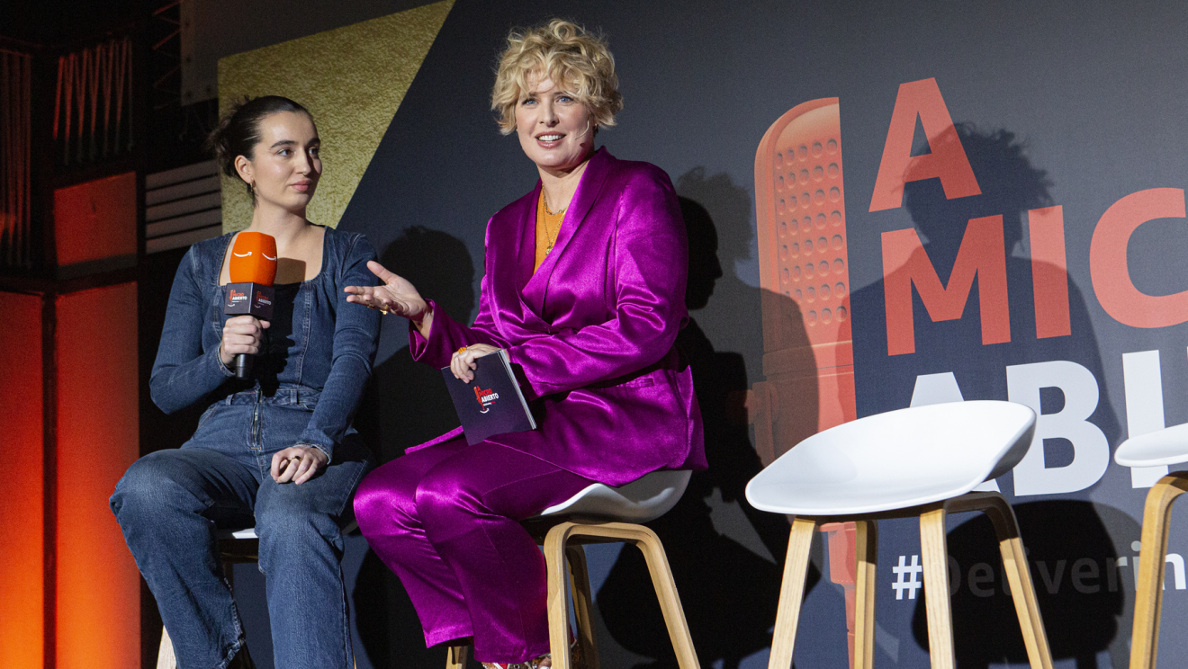 Con el fondo de color azul con el logo de A micro abierto y la etiqueta Delivering Smile, se encuentran dos mujeres sentadas en tabueretes altos. Aparecen la presentadora Tania Llasera (traje chaqueta rosa fucsia, jersey naranja) y otra mujer vestida con mono tejano y con un micro con el logo de Amazon. 