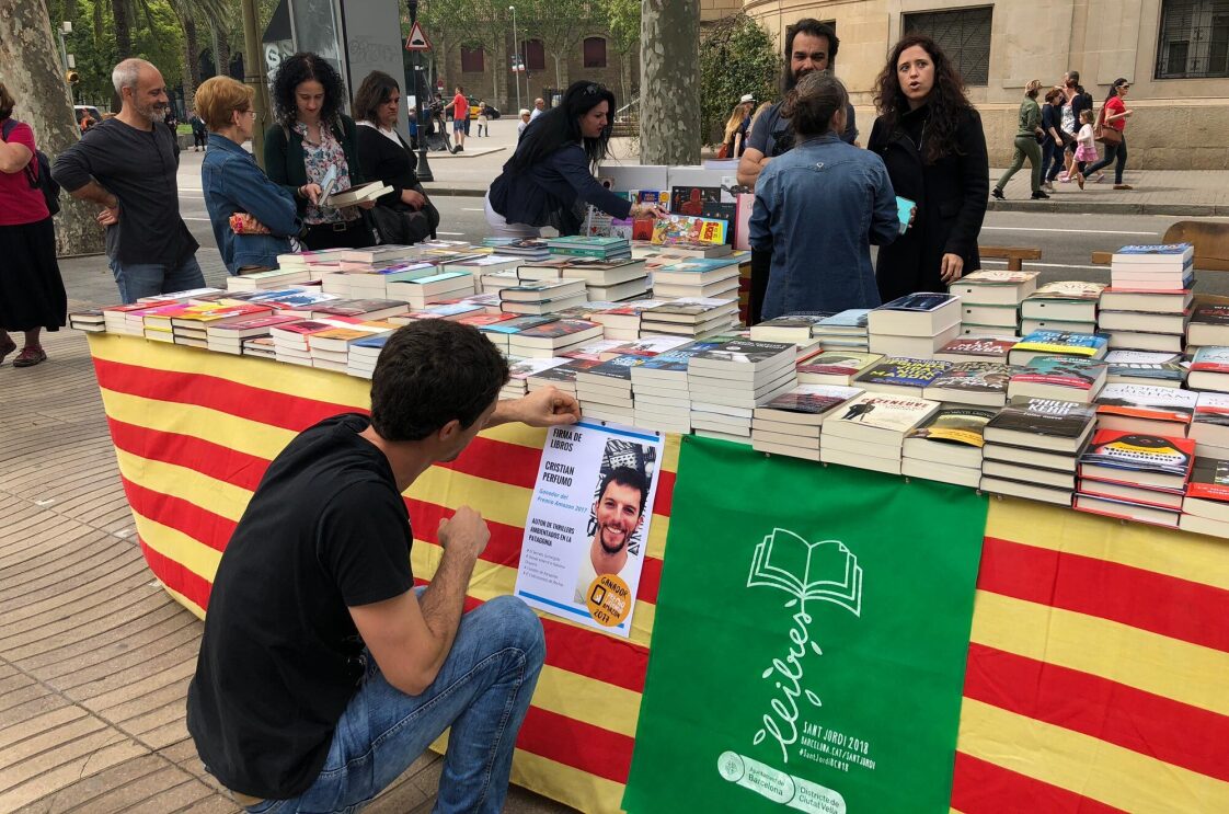 Cristian Perfumo, el escritor de la Patagonia, está de rodillas enganchando un cartel promocional de la firma de sus libros para Sant Jordi en una mesa llena de libros también suyos. 