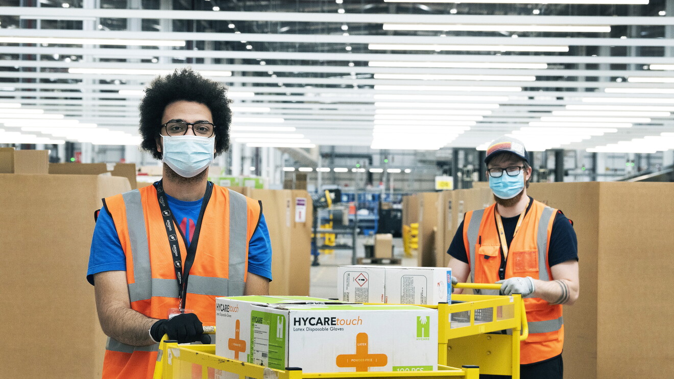 Amazon employees in a fulfilment centre packing medical supplies ready for the government into boxes. They are wearing hi-vis jackets and face masks. 