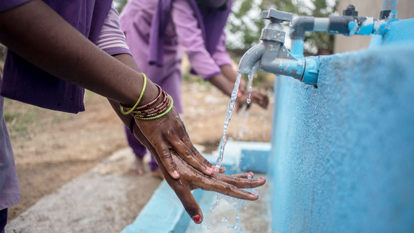 A photo of two people washing their hands at an outdoor faucet. 