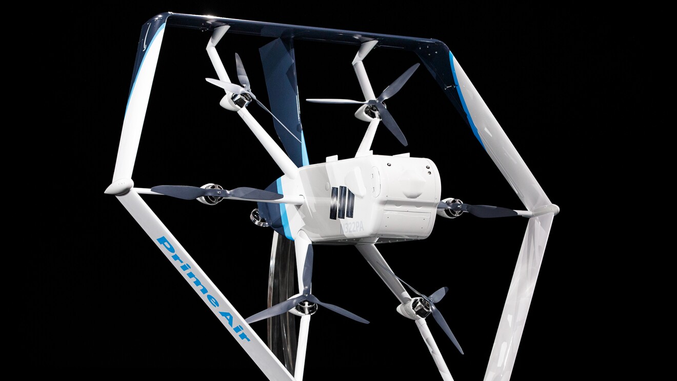 A white and light blue Prime Air drone in front of a black background. Six propellers and a main body are visible in the center of the drone.