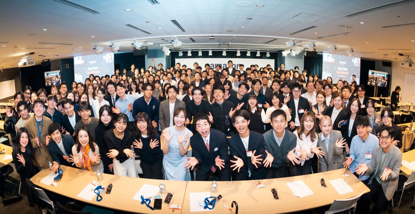 広い室内に集まって集合写真を撮る多数の人々。広げた両手を体の前に出している。魚眼レンズで撮られているので全体が湾曲している。