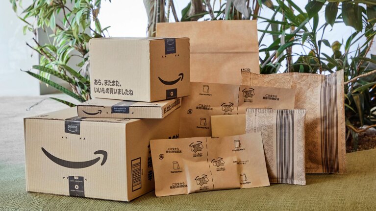 Amazonロゴが入った梱包資材が積まれている。大小のロゴ入り箱や平たい箱、ミシン目が入った大小の紙袋など、全部で9種類ある。背後に観葉植物。