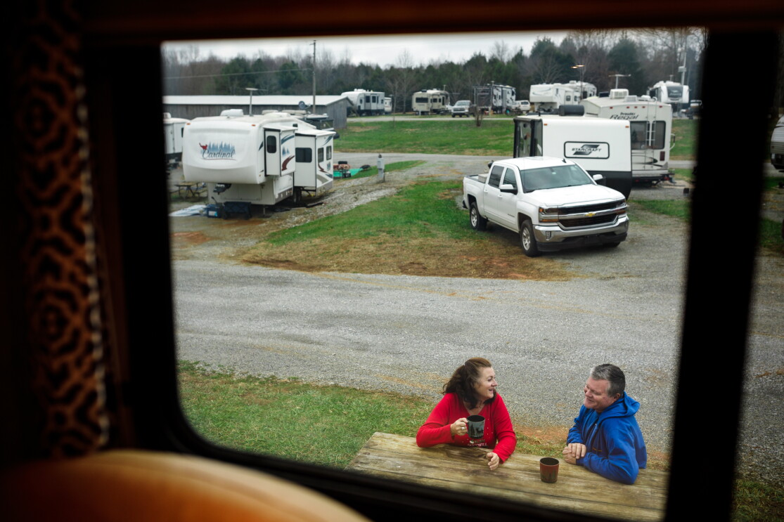 Photographed through a window, a woman and man sit at a picnic bench, drinking coffee.