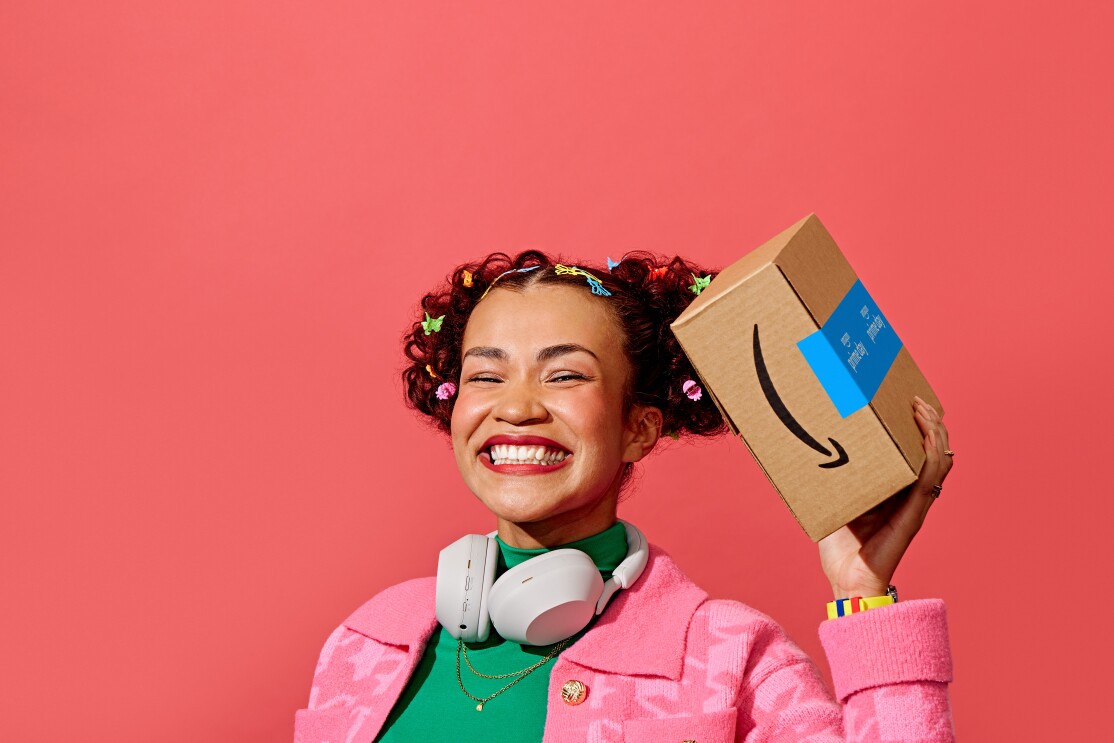 Sobre un fondo rosa aparece una mujer con chaqueta rosa, unos auriculares blancos agarrando una caja de Amazon Prime.