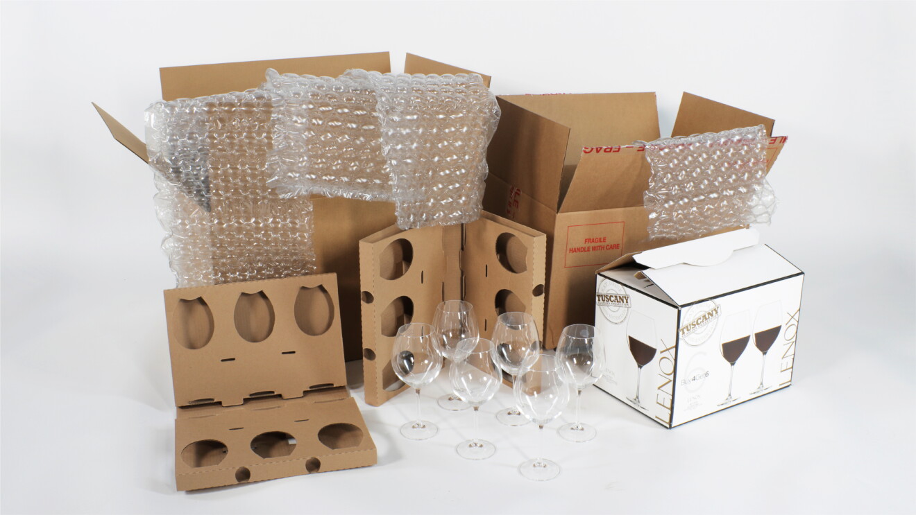 Image of multiple components of stemware packaging before the redesign laid out. Including 6 wine glasses, 2 cardboard inserts, the wine glass packaging, a large cardboard box, 4 sheets of bubble wrap, and a large amazon overbox.