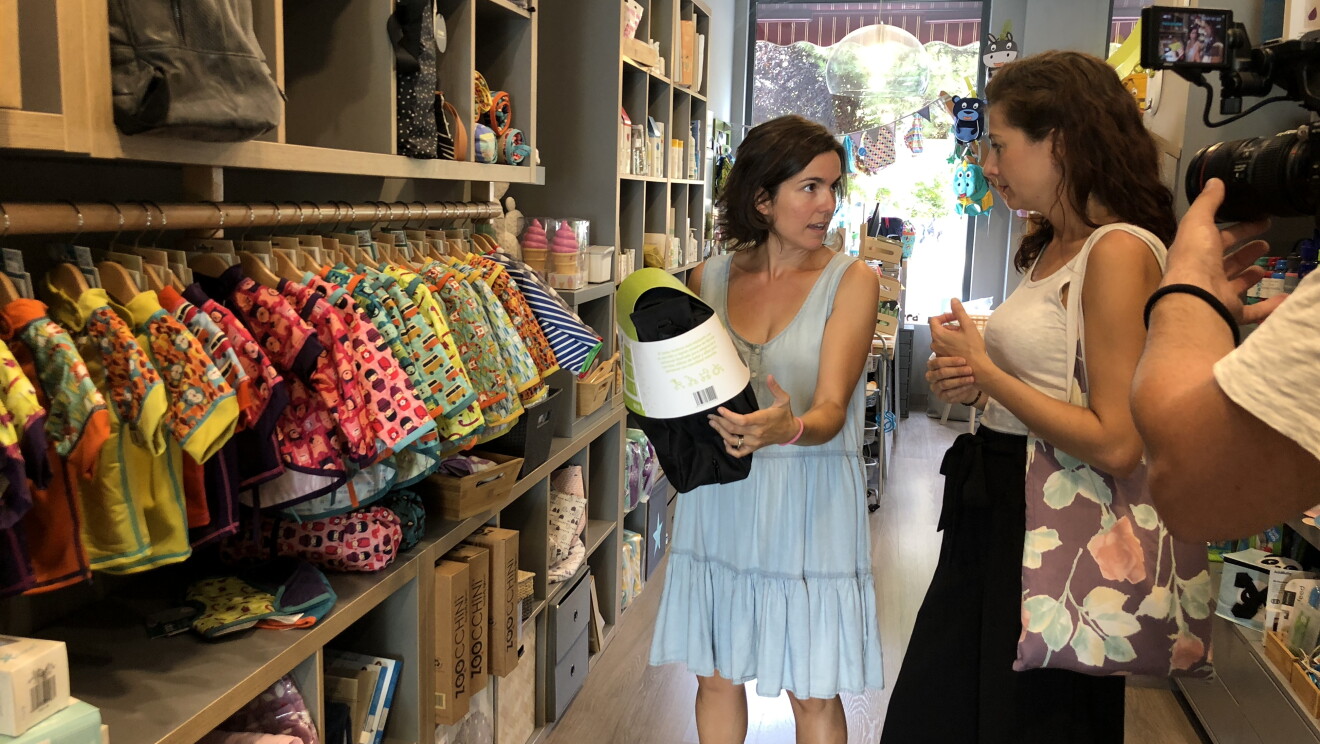 Irene está en una tienda donde se vende su producto. Está conversando con la propietaria de la tienda que tiene en la mano el bolso Andone Dynamic Walks. Irene tiene el pelo largo y viste una camiseta blanca sin mangas y una falda hasta los pies negra. 