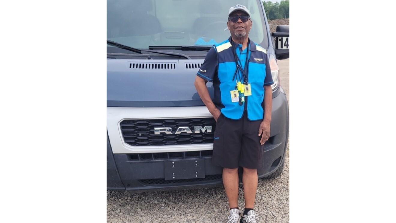 John smiles in front of his Amazon delivery van. He wears his blue Amazon attire with a hat and sunglasses.