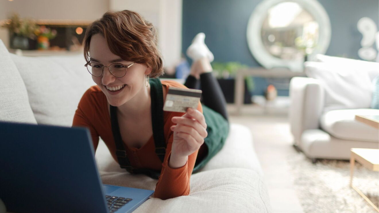 An image of a woman stretched out on a sofa, holding a credit card and smiling in front of a laptop.