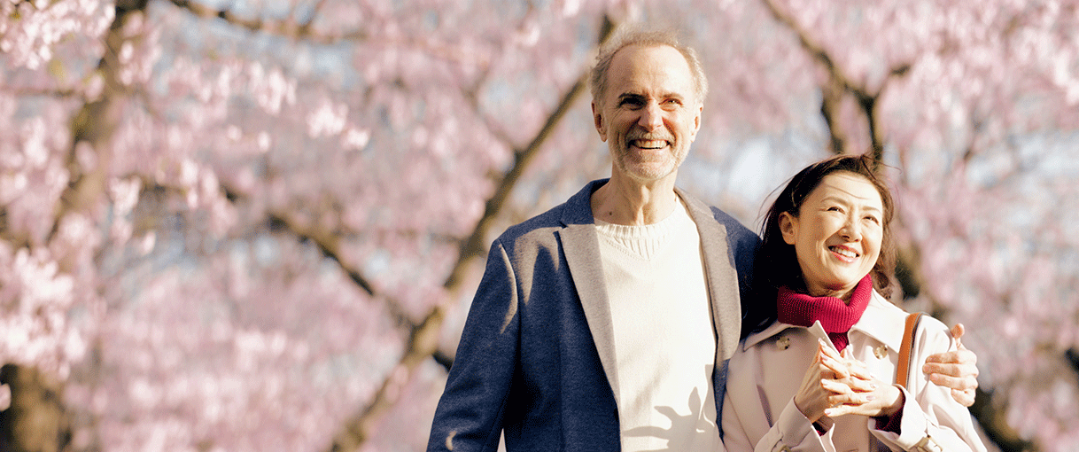 満開の桜の木々を背景に立つ2人の男女。左側の男性が、女性の肩に手を添えている。2人とも笑顔で、女性は体の前で両手の指を交差させている。