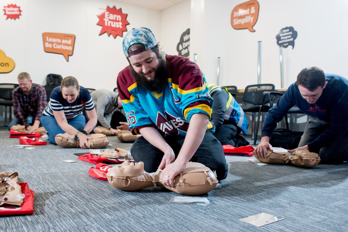 Amazon Operations Supervisor Lloyd Curley kneeling on the floor and practicing CPR on a dummy, along with five of his colleagues doing the same in the background.