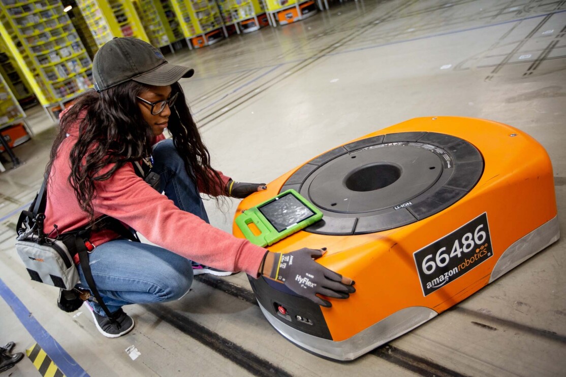 A woman kneels next to a robot, while also working on a tablet, at a Columbus, Ohio fulfillment center, CMH1