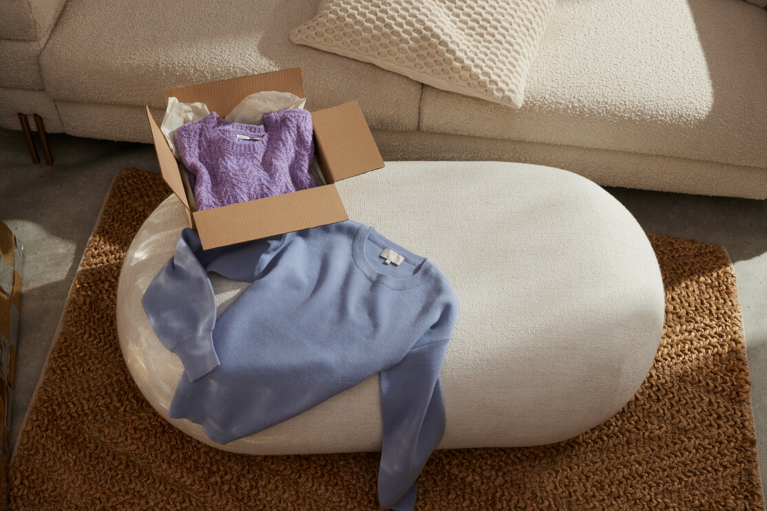 En el salón de una casa hay una sofá con un cojín de color beige, una alfombra trenzada de algodón de yute beige. Encima de la alfombra hay una mesas que es como una pieda y encima una jersei de lana de color azul desplegado y dentro de una caja de color marrón, otro jersey trenzado de color morado.