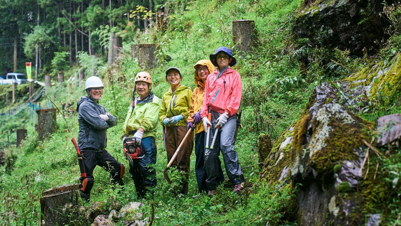 森の斜面に立つ作業着姿の5人の人物。ヘルメットを着用、チェーンソーや植木用の鋏を持っている。斜面には切り株が多数。