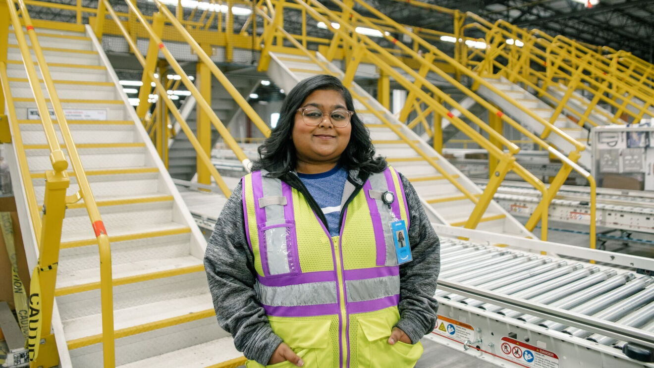 Amazon employee stands smiling with her hands in her pocket in front of a conveyor built in a fulfillment center.