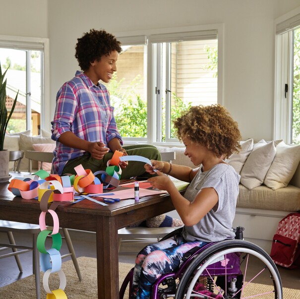 A mother sits on a dining room table as she and her daughter laugh, while making a paper chain. Behind them, Amazon Echo is on a console table.