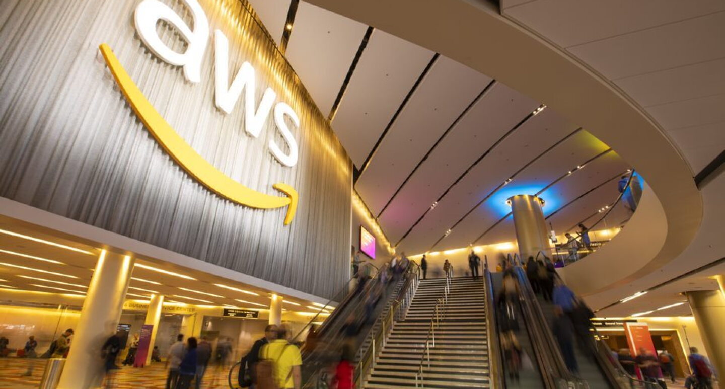 Edificio_AWS. Entrada al edificio de AWS de Amazon. En la entrada con el suelo gris y rombos de color rojo, naranja y amarill. Hay cuatro escaleras automáticas y unas escaleras en medio. El texto es blanco y en una columna del fondo hay luz indirecta azul. Hay gente subiendo y bajando pero difuminada.  