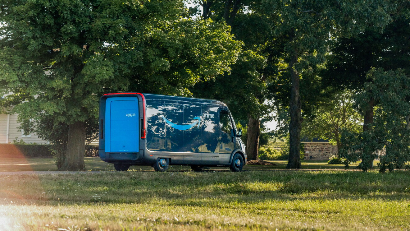 En una carretera que passa entre árboles se ve al fondo una furgoneta Rivian con el logo de Amazon y la palabra Prime. La furgoneta tiene dos tonos azules, más y menos intenso. 
