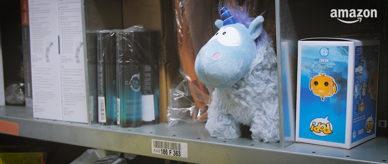 Ein blaues Plüsch-Einhorn steht in einem Lagerregal, zwischen diversen Artikeln.