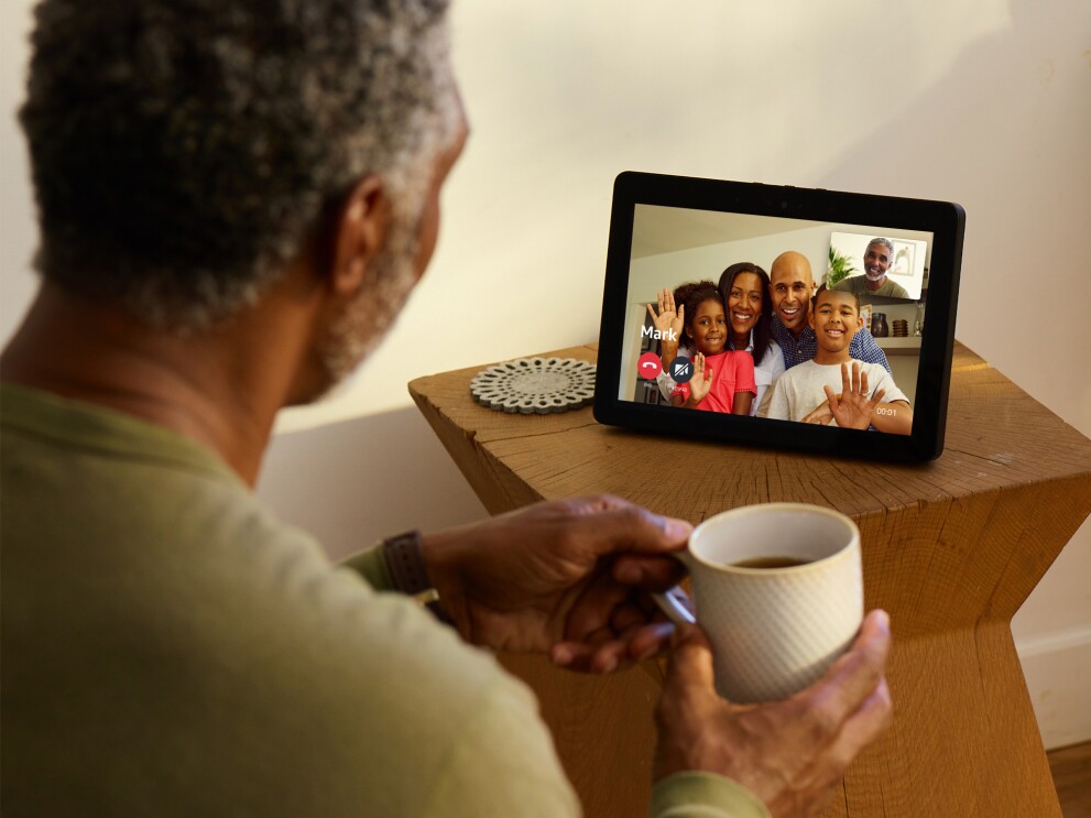 An Echo Show device on a wooden side table. On the screen, a family of four are smiling and waving on the screen. Facing the device is a man, holding a coffee cup. The screen shows his smiling face. 