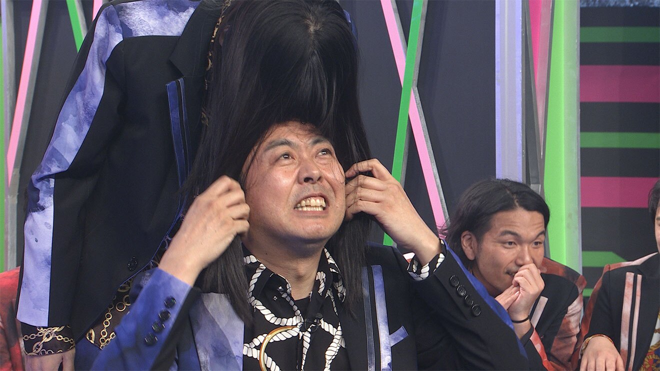 黒地に黄緑とピンクの装飾がある壁を背景に、派手な衣装の男性たち。後ろに立った長髪の人物が前にかがんで、前に座っている男性の顔に髪の毛をかぶせている。右側にも2人の人物が座っている。