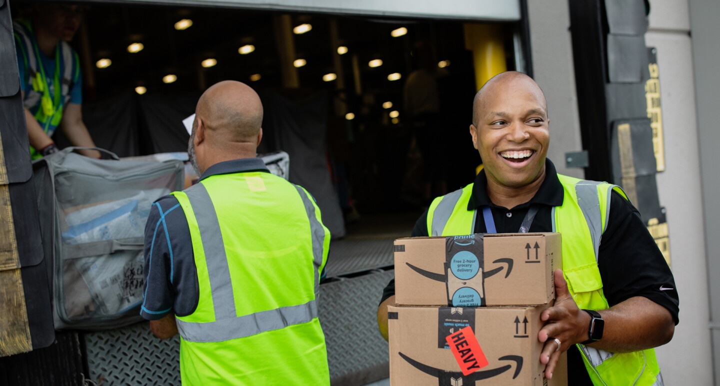 A smiling man holds boxes while people in the background load a truck. 
