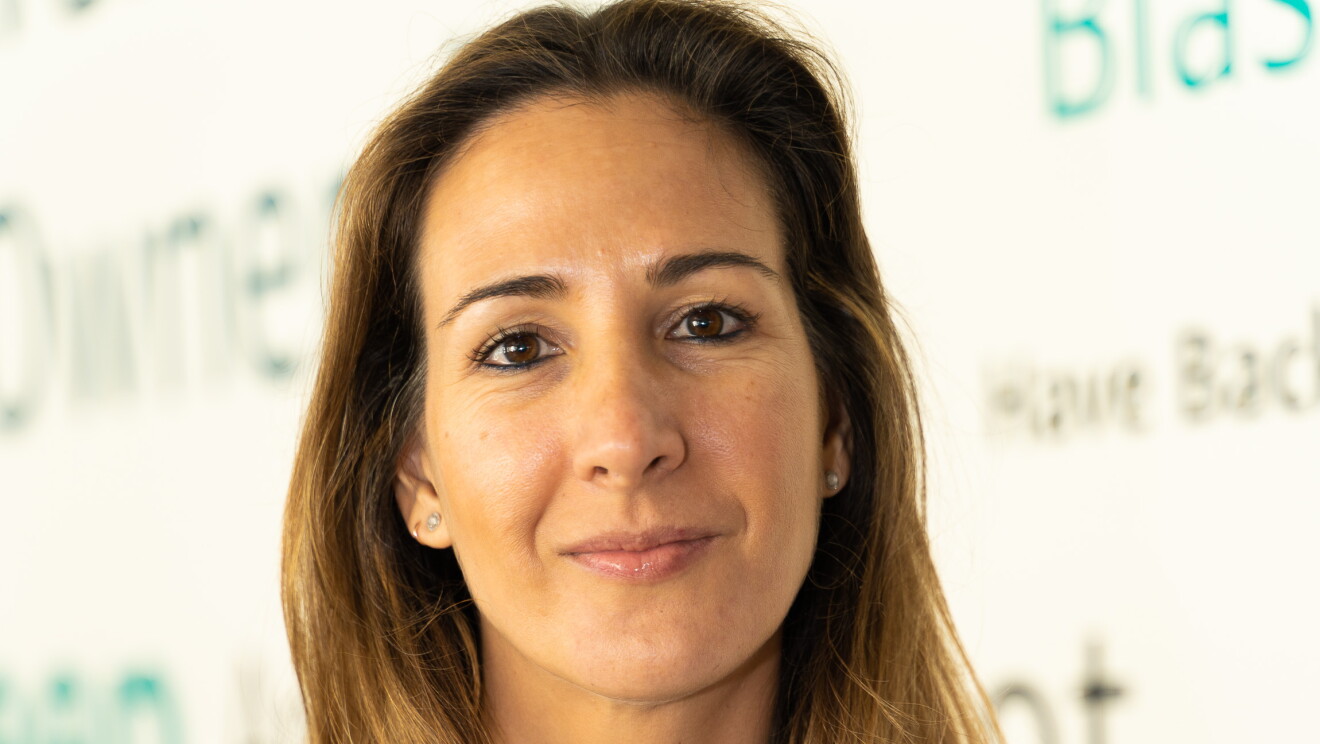 Silvia Cubero, 3P Program Manager, Marketplace Recruit and Dev. mirando a cámara y con el ordenador en las manos. Con una camisa blanca, dos collares, pendientes y el pelo por debajo del hombro. 
