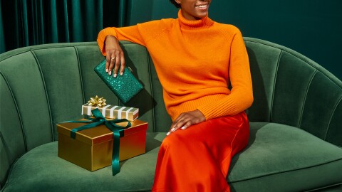 A woman sits on a green couch smiling. She is holding a sparkly, green clutch purse and there are gift boxes around her.