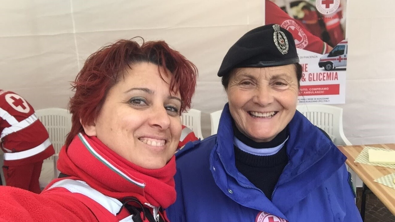 Due volontarie della croce rossa si scattano un selfie. La donna a sinistra è vestita di rosso e regge il telefono mentre scatta la foto, la donna a destra è vestita di blu e indossa un basco. 
