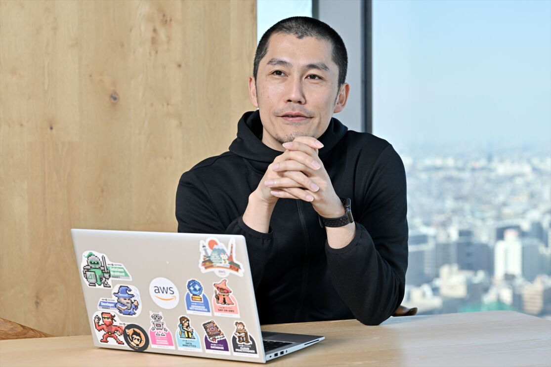 黒いジャケットを着た髪がない男が指を絡ませている。室橋弘和さん。彼の前にはたくさんのスタンプが貼られたPCが置かれている。