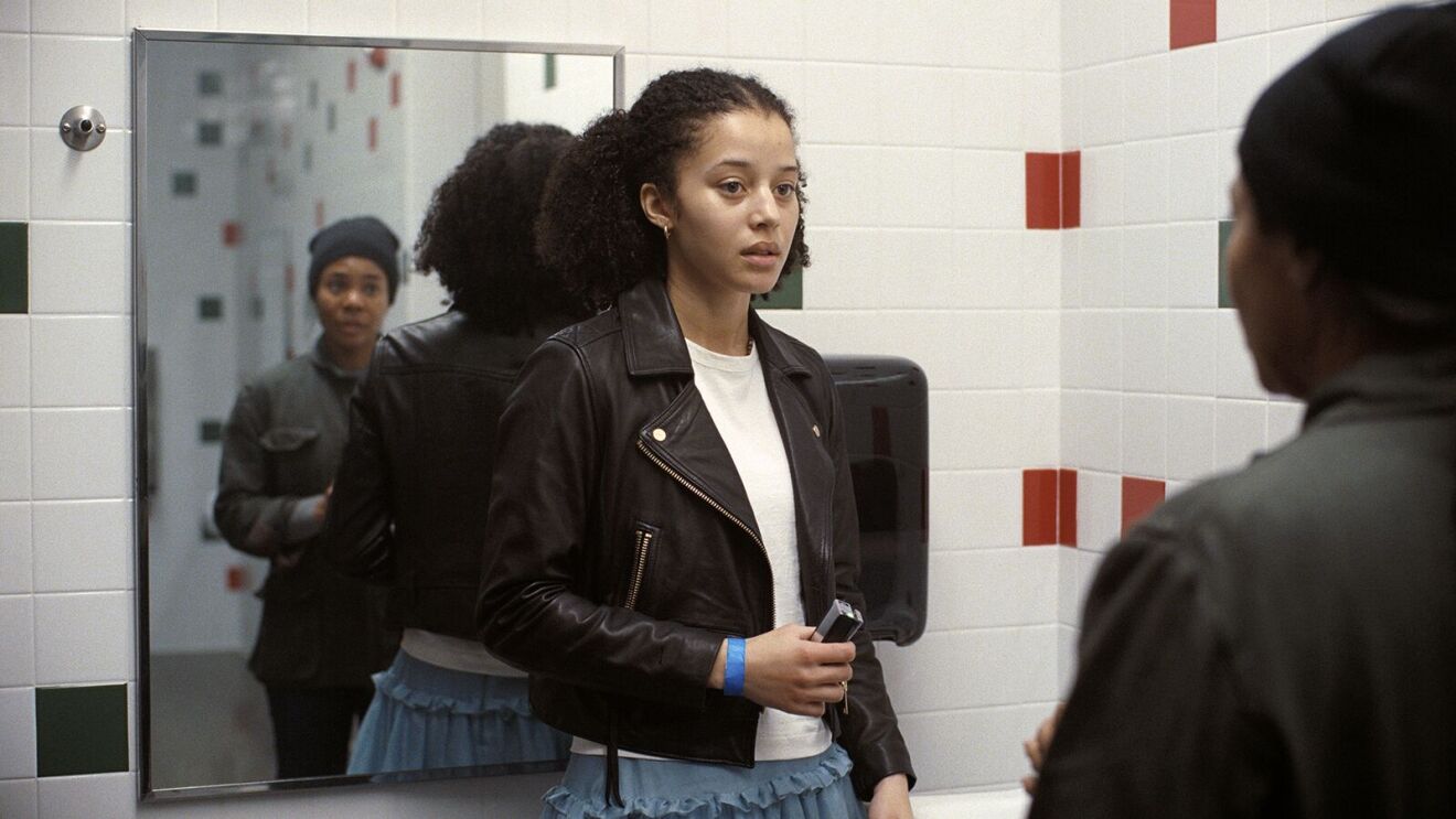 Willa Ferguson (Chase Infiniti) in leather jacket stands in tiled bathroom, speaking with Deandra (Regina Hall)