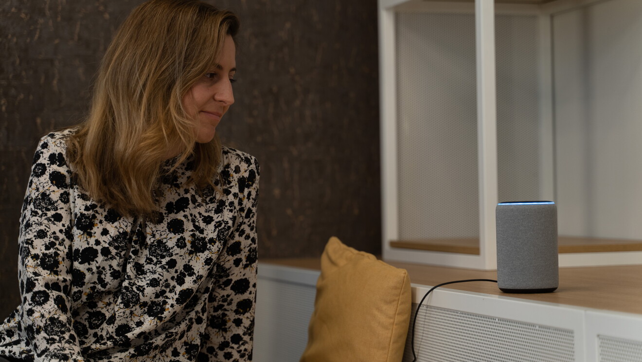Marta Morales, Senior Product Manager de Alexa. Con una camisa estampada y el pelo castaño claro, Marta mira fijamente un dispositivo Echo de color gris colocado sobre un mueble en la parte derecha de la imagen.