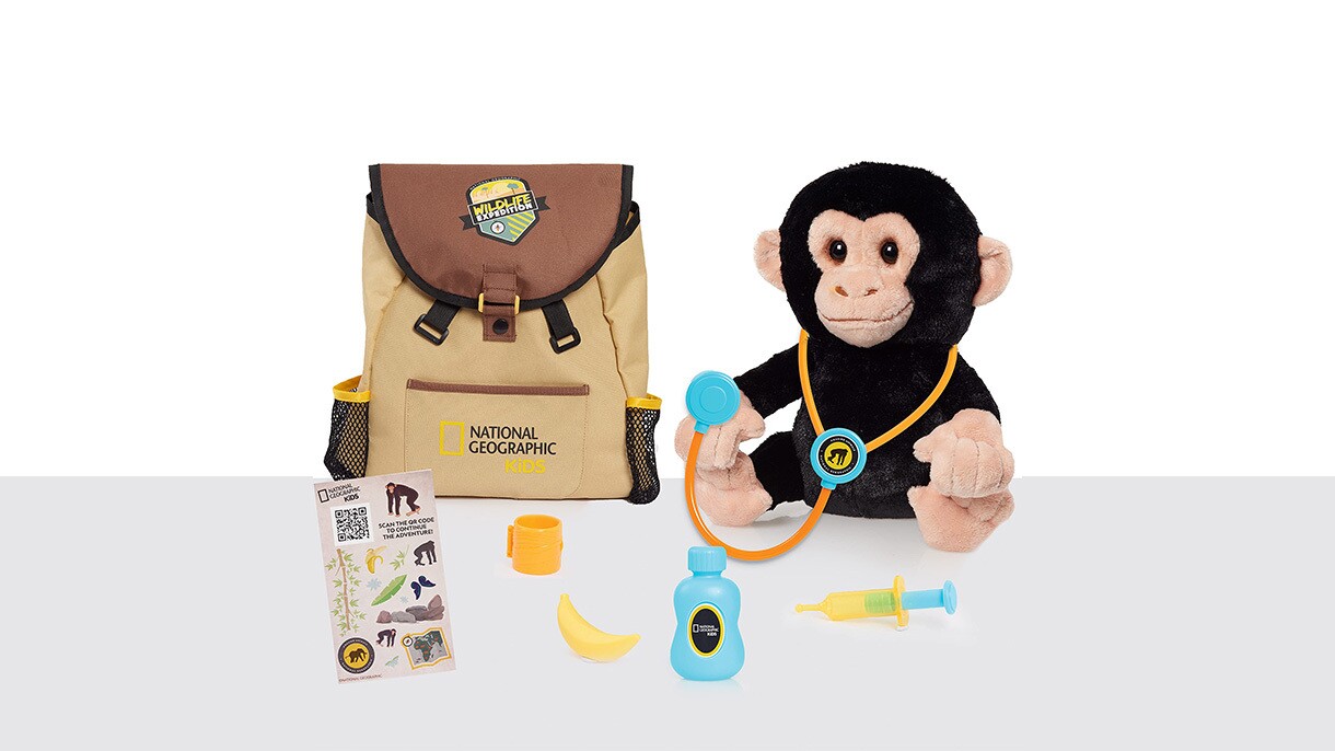 An image of a brown backpack with a stuffed monkey toy next to it. The monkey toy has a stethoscope on its ears. There are other accessories like a bottle and a banana in front of the monkey and the backpack.