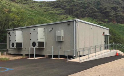 A gray prefabricated equipment shelter/building with multiple exterior HVAC and ventilation units mounted on its wall, set on a paved pad with a metal access ramp and railing. A steep green vegetated hillside rises directly behind it.