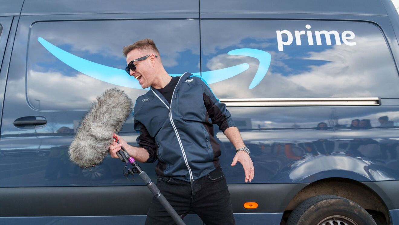 Westlife's Nicky Byrne, wearing an Amazon-branded jacket, stands in front of a Amazon delivery van. He is pretending to sing into a boom mic