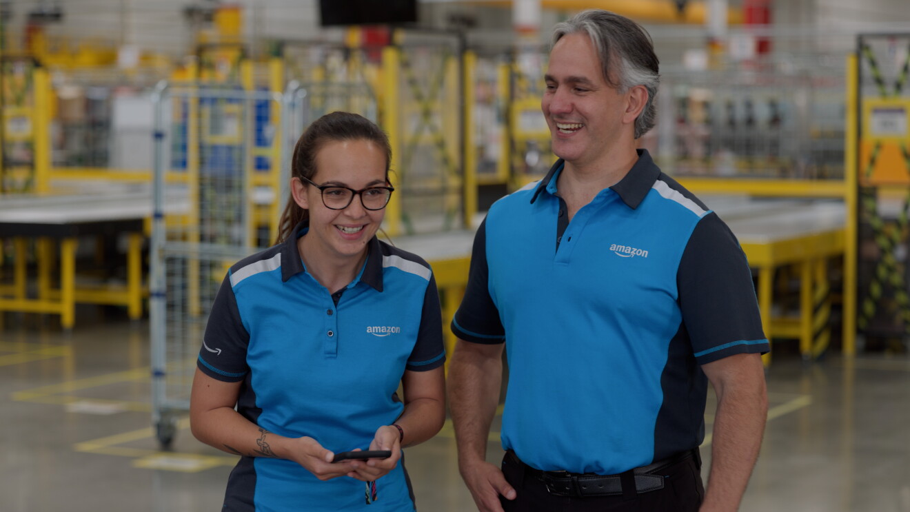 María y Jonathan de Dolinski - Becerra Services, S.L. dentro de un centro logístico de Amazon. Los dos van con el mismo polo con franjas de diferentes tonalidades azules y el logo de Amazon en la manga y en el pecho. Los dos se están riendo. Jonathan tiene el pelo corto canoso y tiene las manos en los bolsillos. María lleva el pelo largo pero recogido en una coleta, unas gafas de pasta marrones, tiene pecas en la cara y está agarrando un móvil. 

