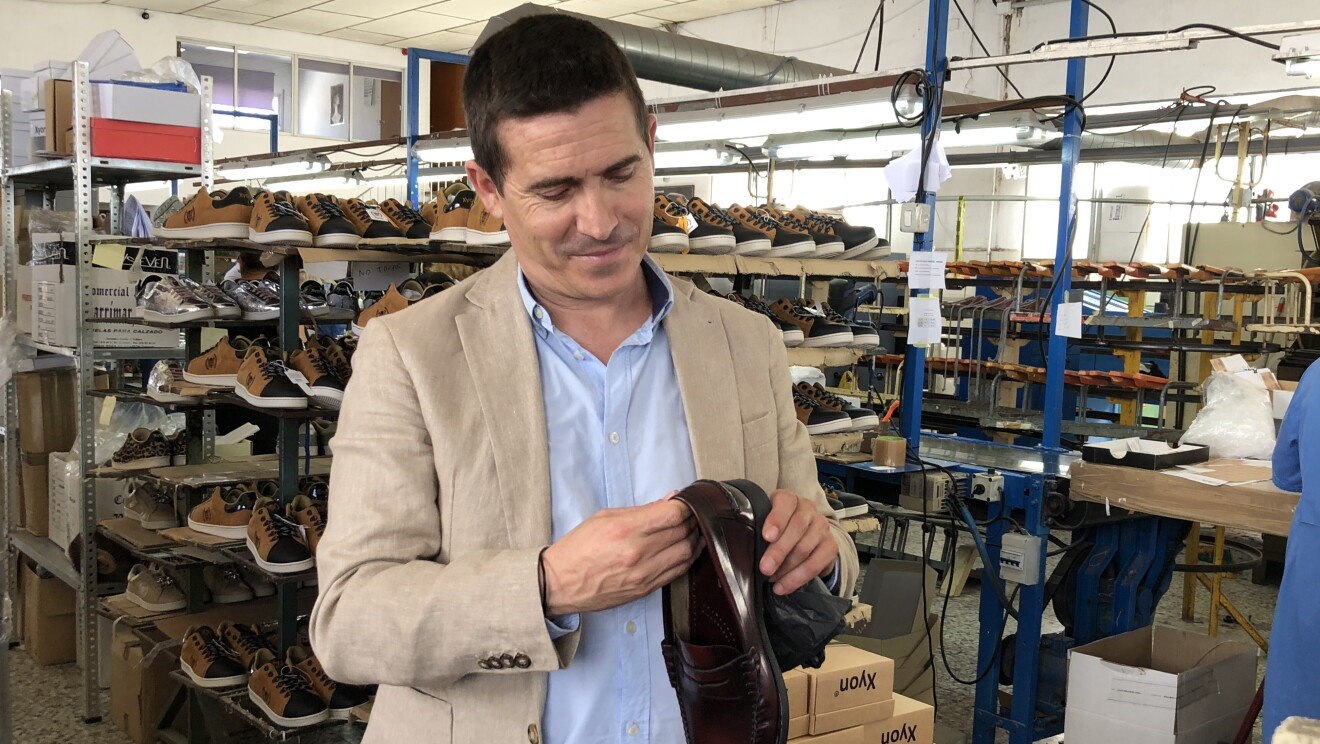 José Antonio Mollá, fundador de Castellanisimos, en una de sus fábricas, observando un zapato castellano que tiene en sus mannos. Detrás hay cahas de zapatos y una persona trabajando en una mesa. 