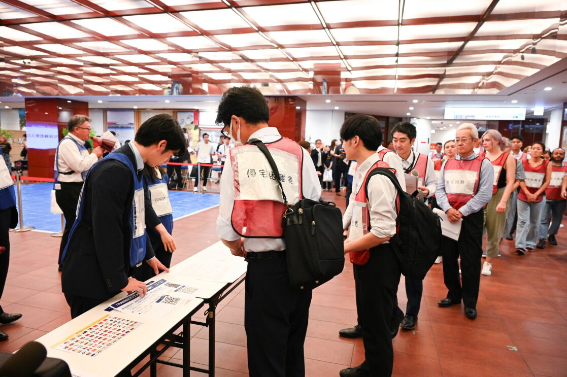 「帰宅困難者」という表示がつけられているオレンジベストにを着ている多数の人が長テーブルに設けられた受付の前に並んでいる。長テーブルにはスーツに青いベストを着ている男性2人が紙に書類を指で示している