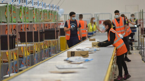 Un rullo portapacchi in un magazzino Amazon. Scatole si alternano a buste in carta. Si vedono in secondo piano alcuni dipendenti del magazzino. 
